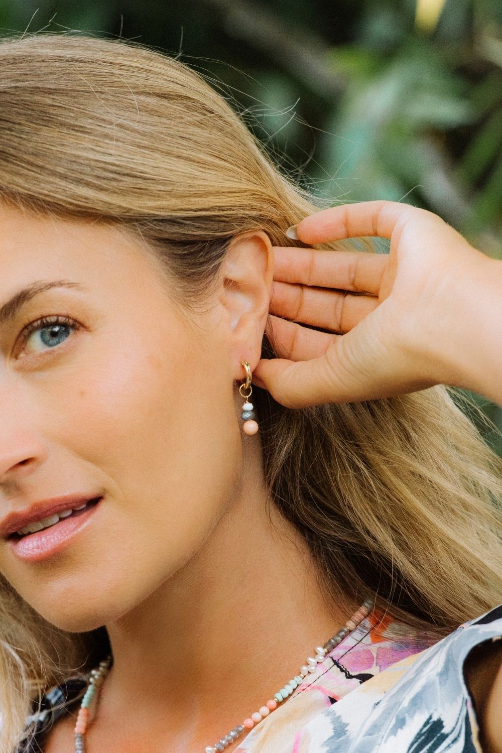 Woman adjusting a gold huggie earring with a coral cove charm and a blurred green background