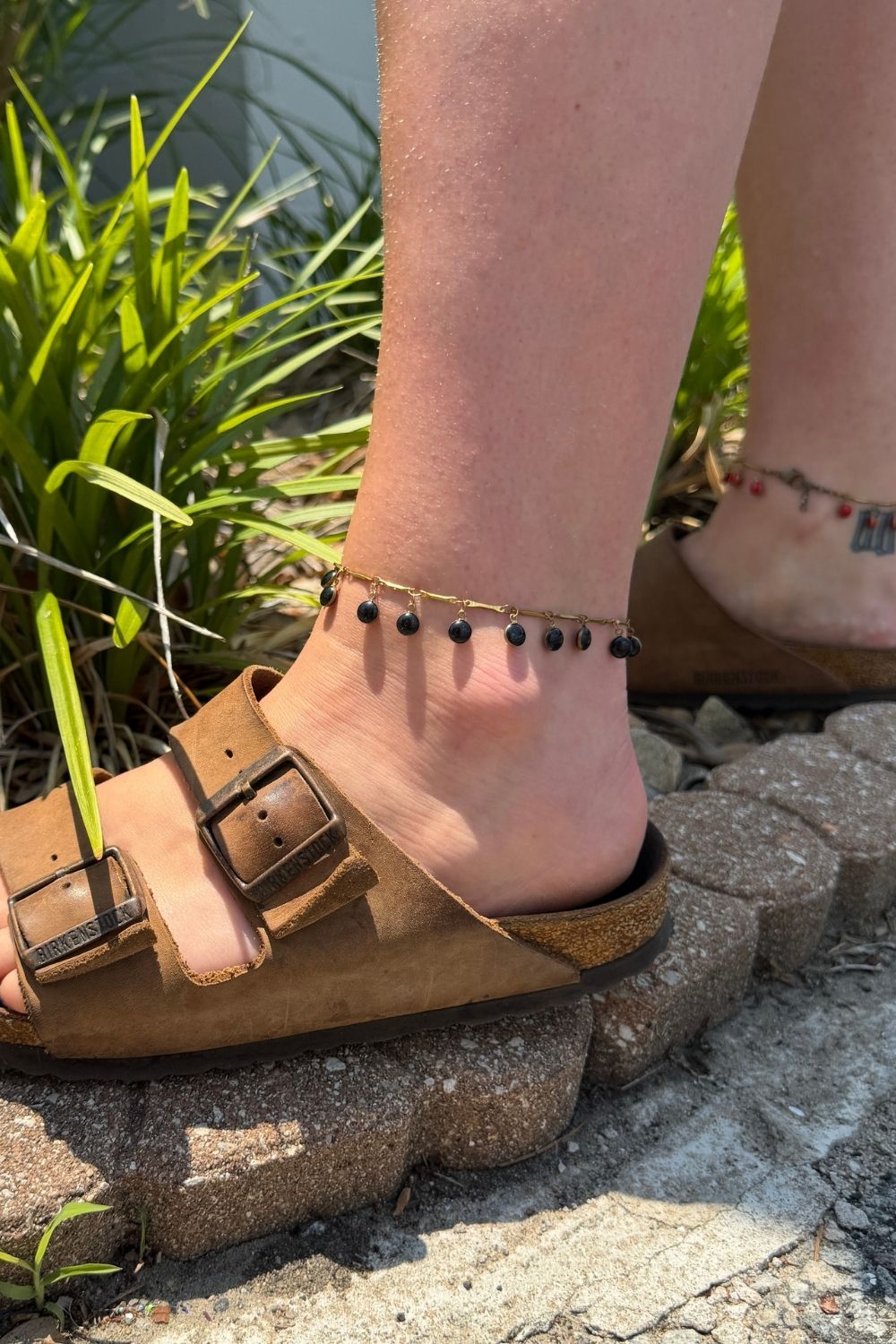 wearing a dancing dreamer black anklets standing on a stone surface with greenery in the background.