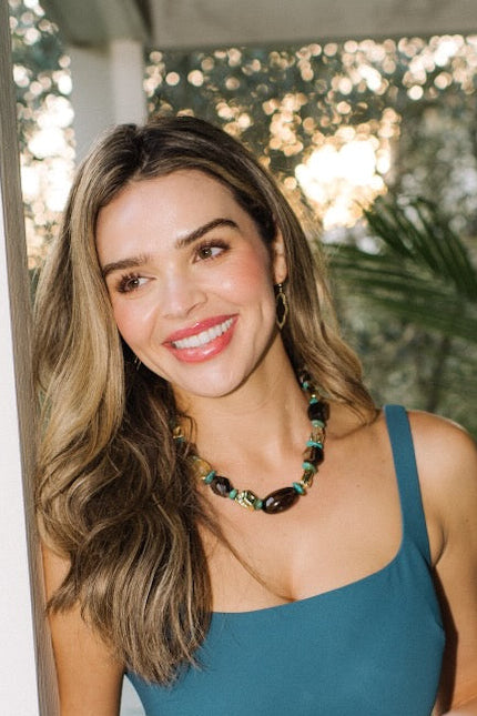 Woman in a teal dress holding a silver gift box on a porch with greenery in the background