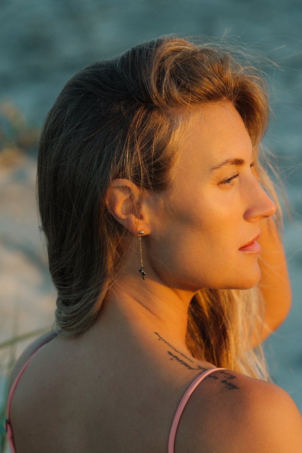 Woman wearing riptide shark tooth earring and tattoos on her shoulder, looking towards the water.