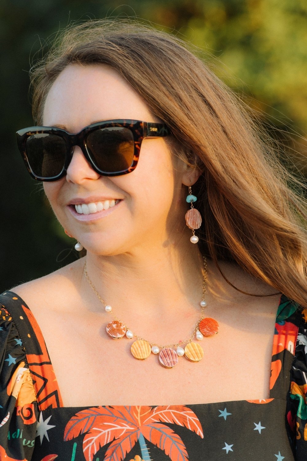 Woman wearing sunglasses and a colorful Scallop Sunset necklace with shell pendants and pearl accents.