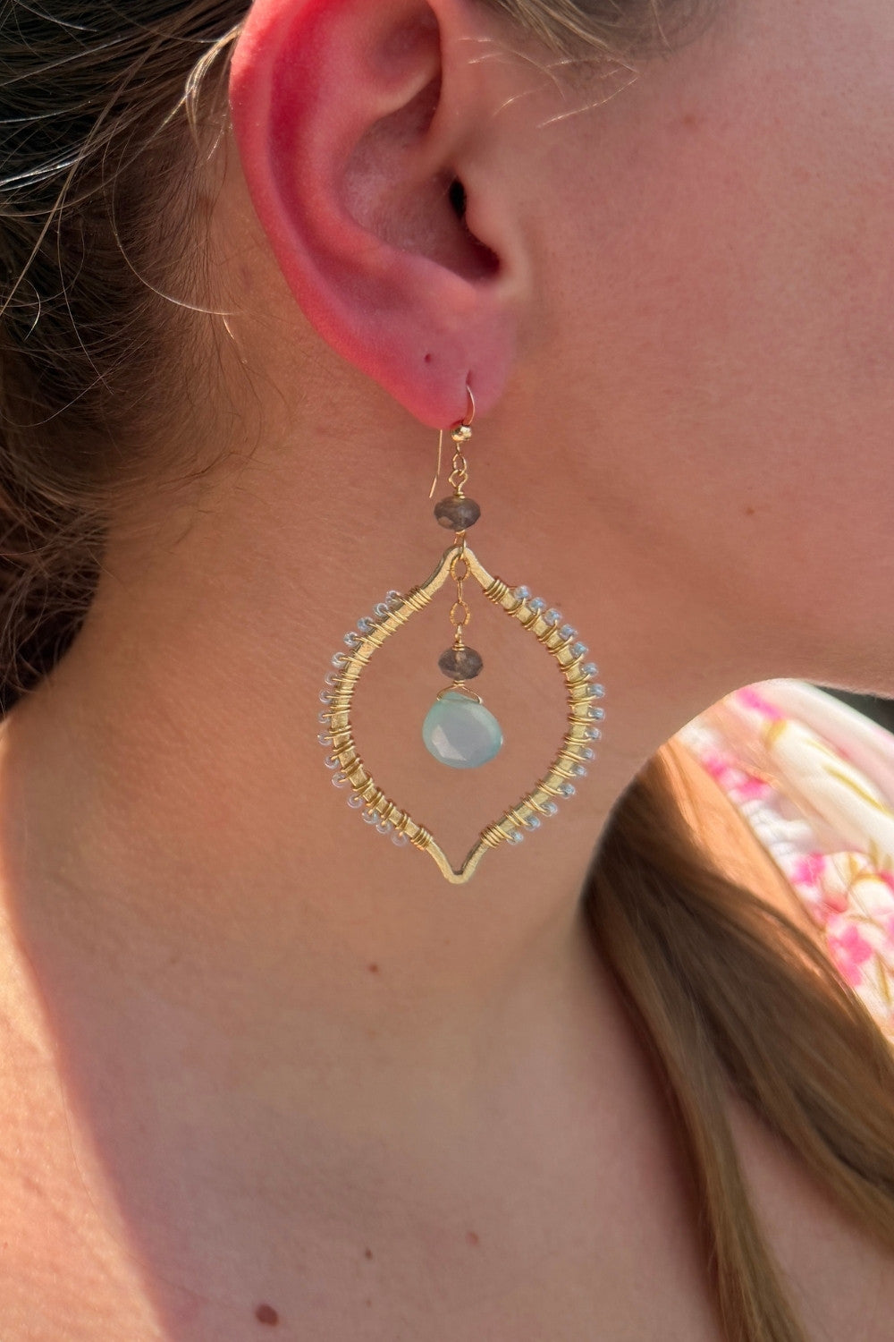 Close-up of a person wearing a gold solstice earring with a blue chalcedony gemstone.