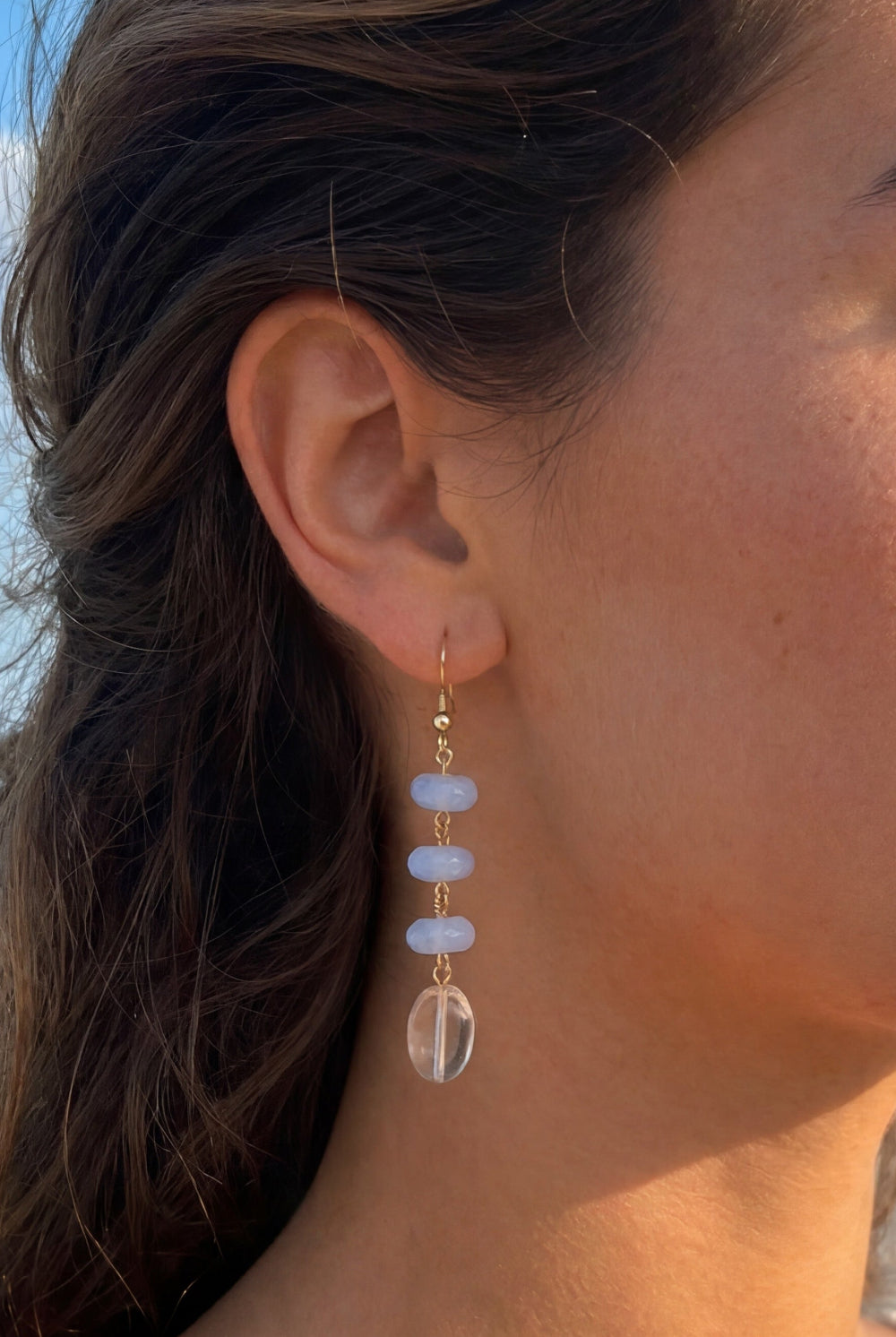 Woman on a beach wearing earrings with a clear stone and blue gem.