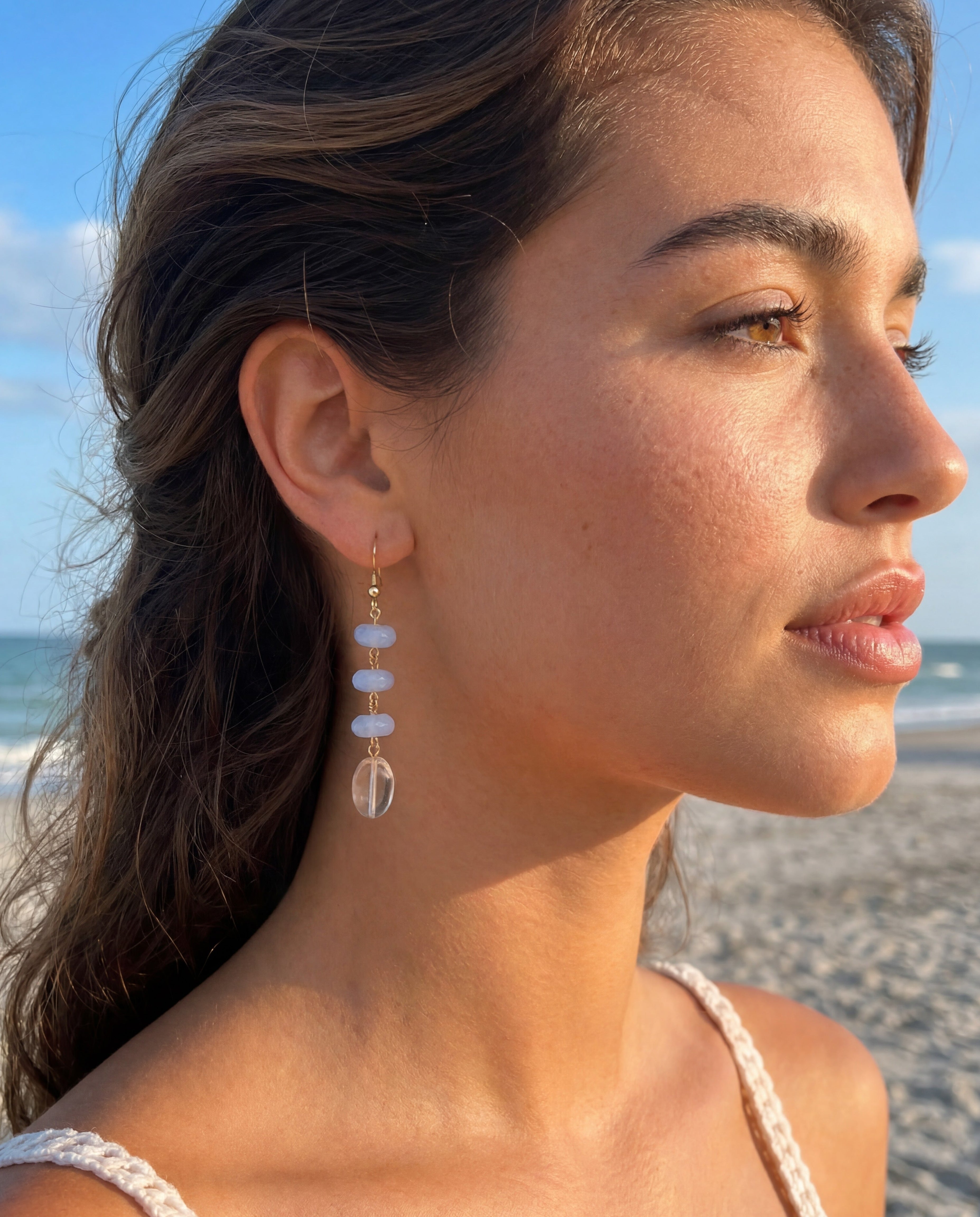 Woman on a beach wearing earrings with a clear stone and blue gem.