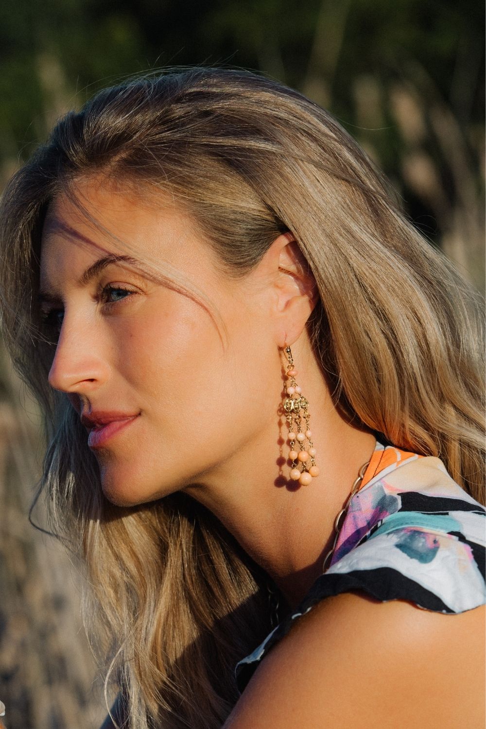 Woman wearing large, dangling coral chandelier earrings with a blurred natural background