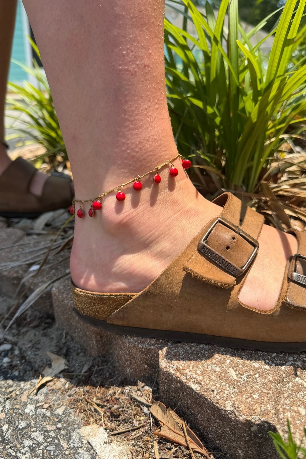 wearing a dancing dreamer red anklet, standing on a stone surface with green plants in the background.