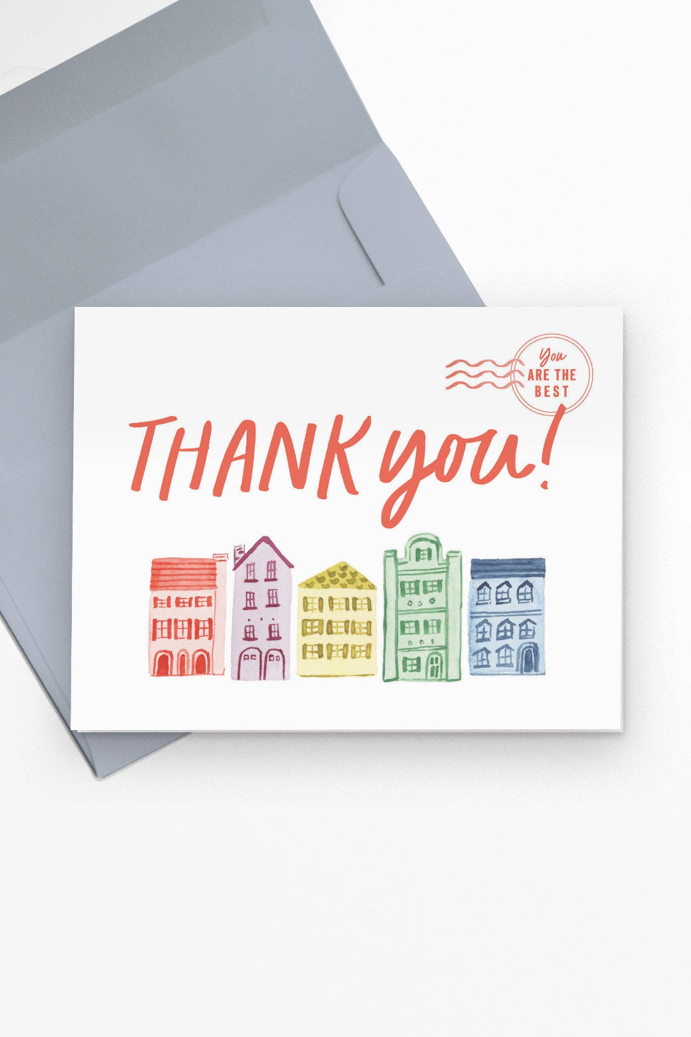 A white greeting card with the words “THANK you!” in coral orange and a row of colorful illustrated buildings along the bottom, shown with a gray envelope on a white background.