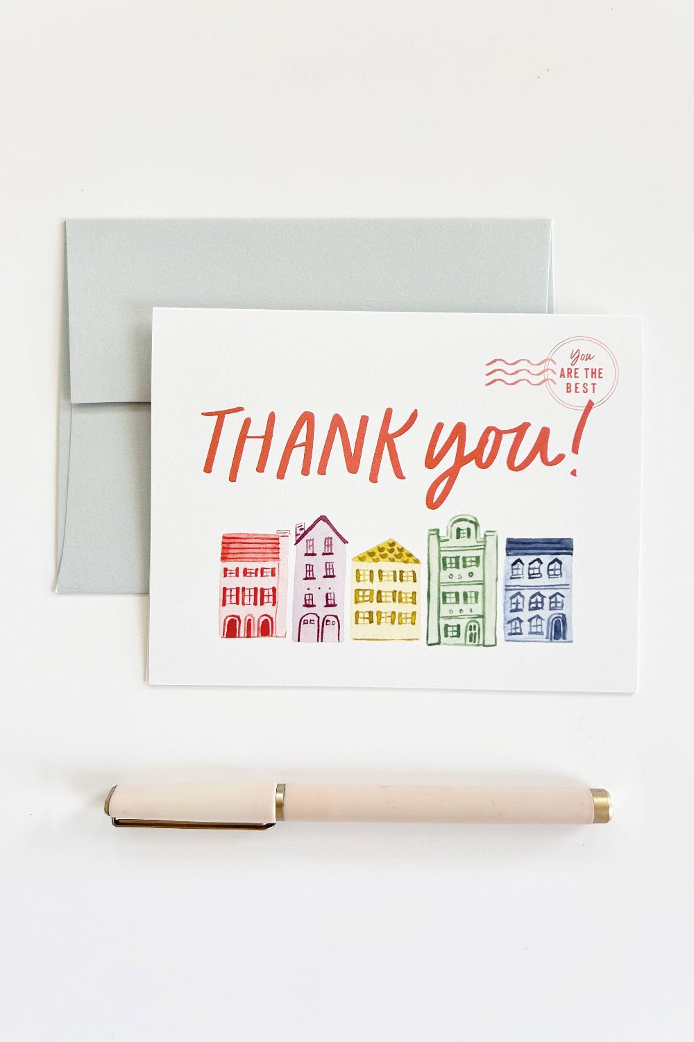 A white thank-you card with colorful illustrated buildings and the words 'THANK you!' in coral script, shown with a light gray envelope behind it and a pale pink pen on a white surface.