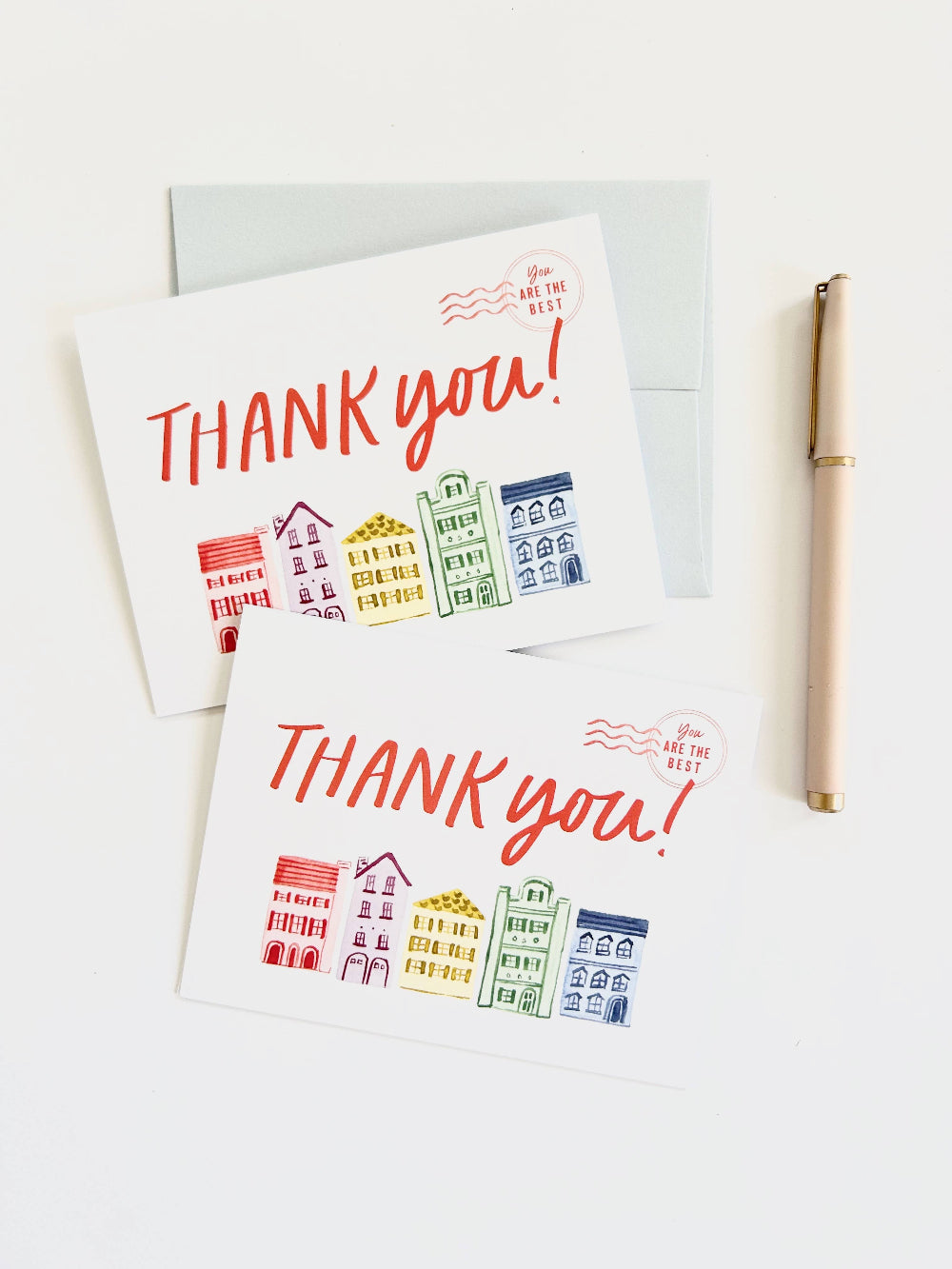 Two white thank-you cards with colorful illustrated buildings and red 'THANK you!' text, arranged on a white background with a light blue envelope behind them and a gold pen to the right.