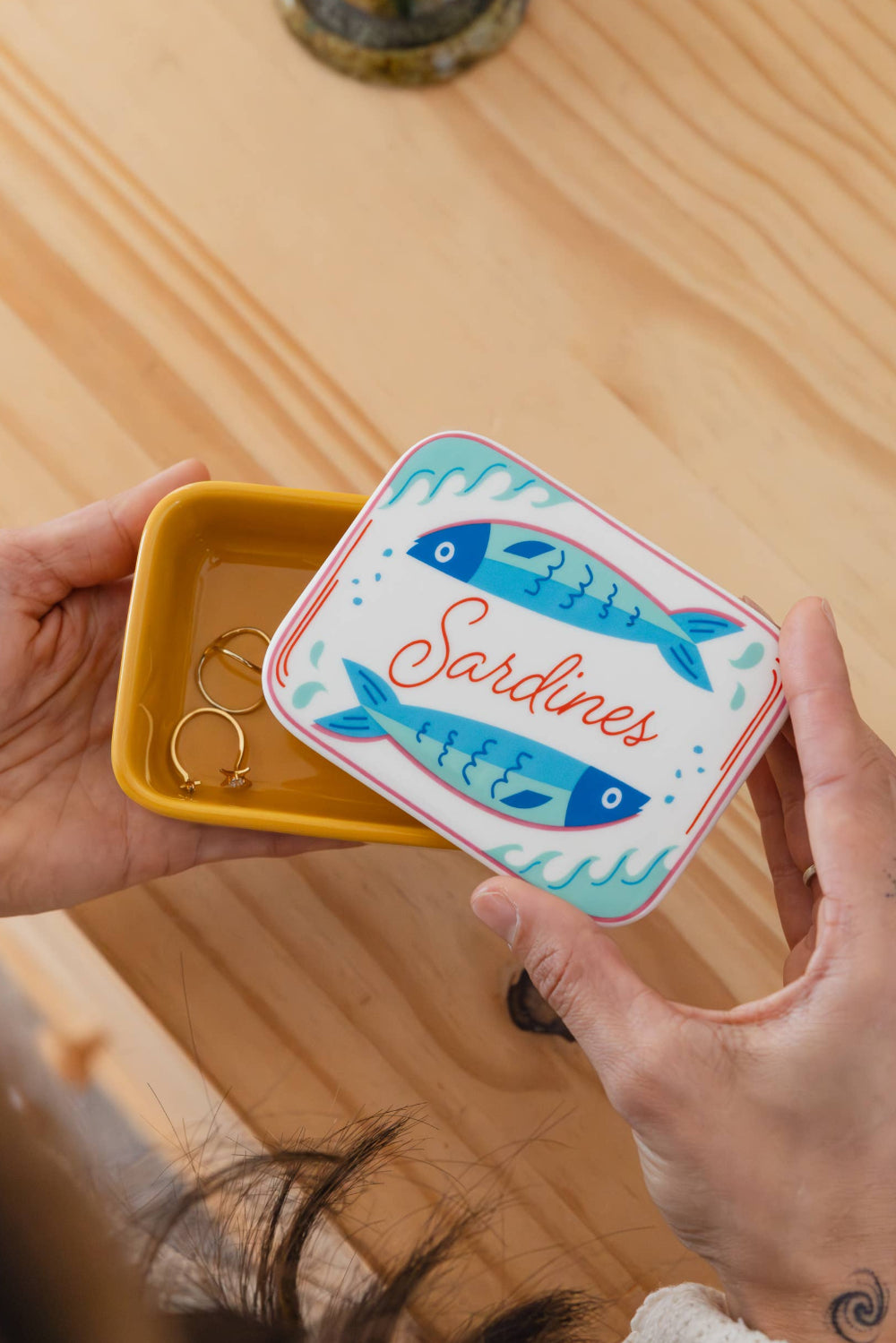 Person holding a small yellow ceramic box with a decorative lid featuring fish illustrations and the word 'Sardines'.