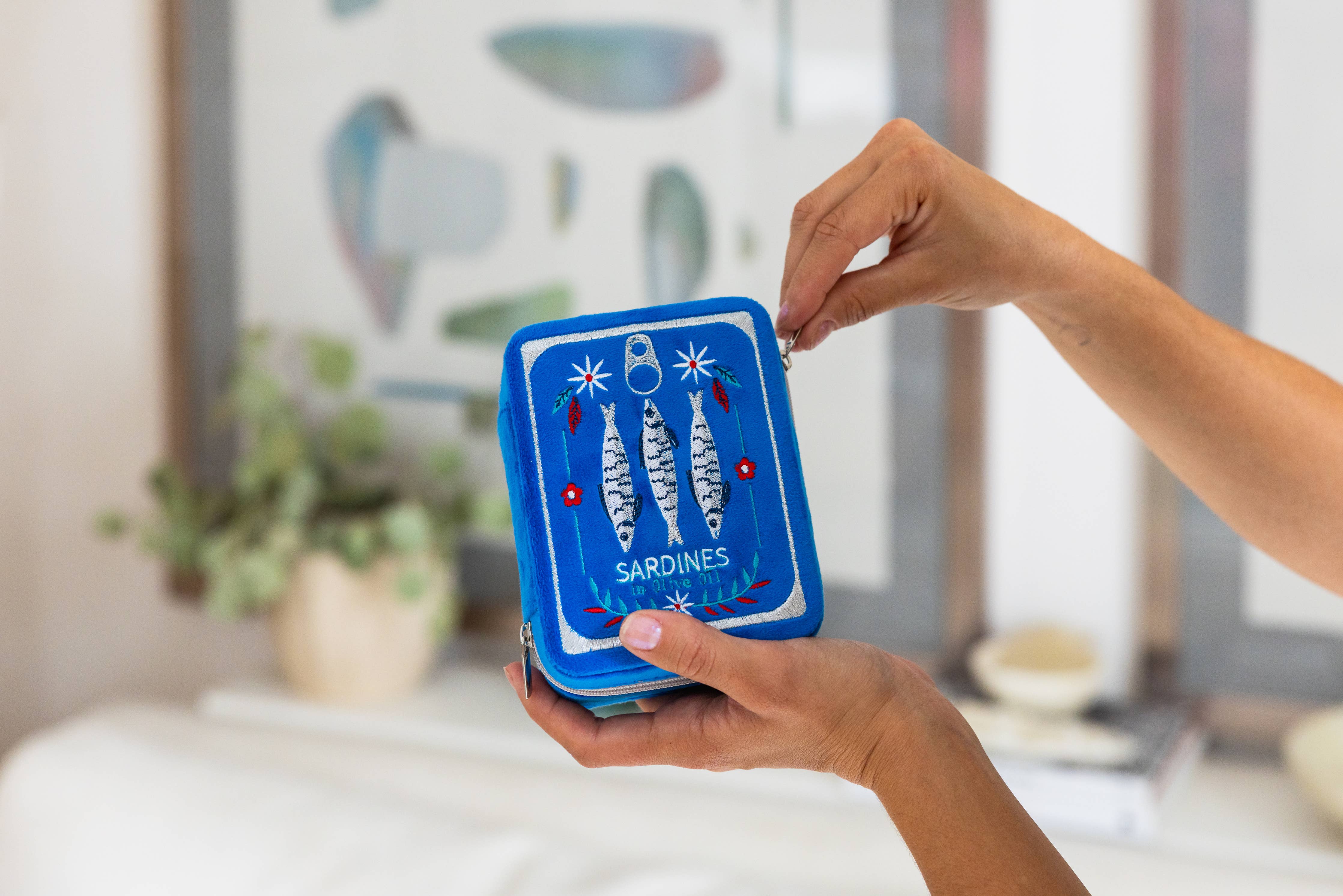 Hand holding a blue jewelry case with fish illustration and text, blurred indoor background