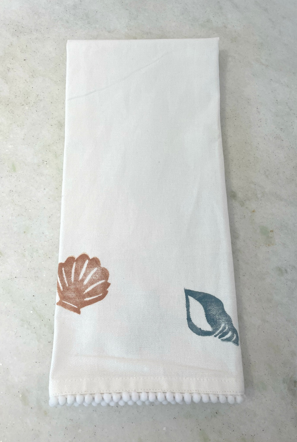 A linen tea towel with a pom-pom trim, featuring printed seashell designs in terracotta and slate blue, laid flat on a light stone surface.