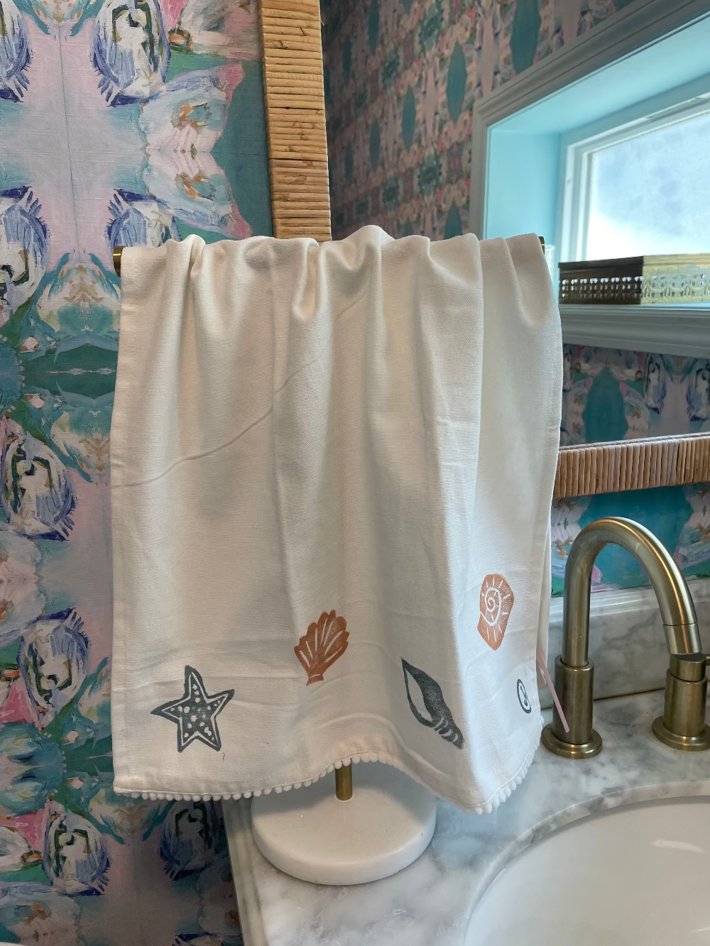 A linen tea towel with hand stamped seashell motifs hanging on a stand in a bathroom next to a sink and brass faucet.