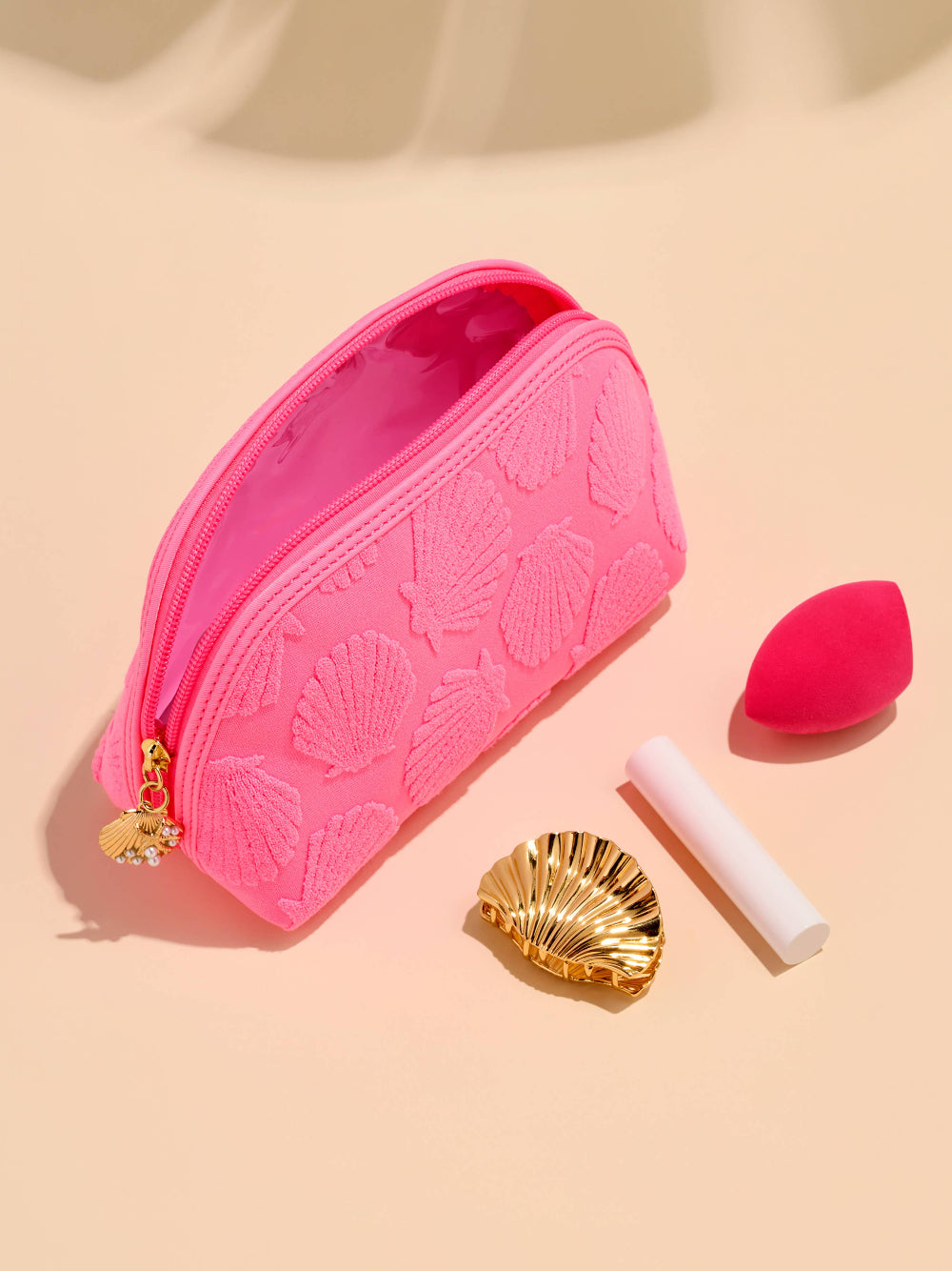 Pink makeup bag with shell design, a red beauty blender, and a gold shell clip on a beige background.