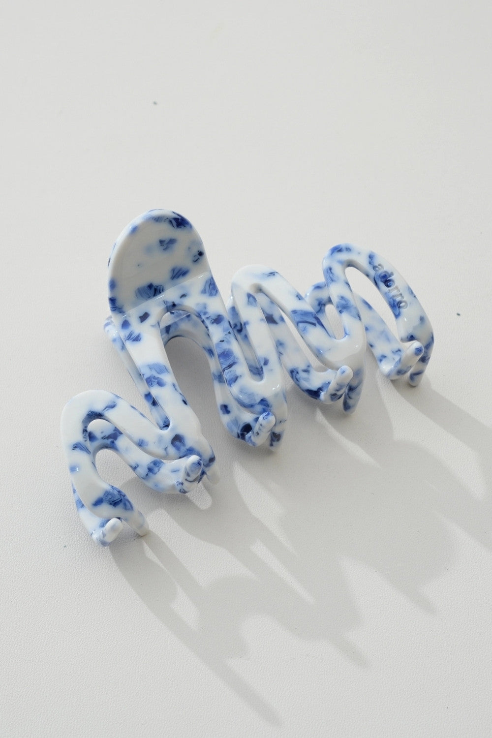 A single wavy claw-style hair clip in a white and blue marbled pattern, photographed on a clean white background with soft shadows.