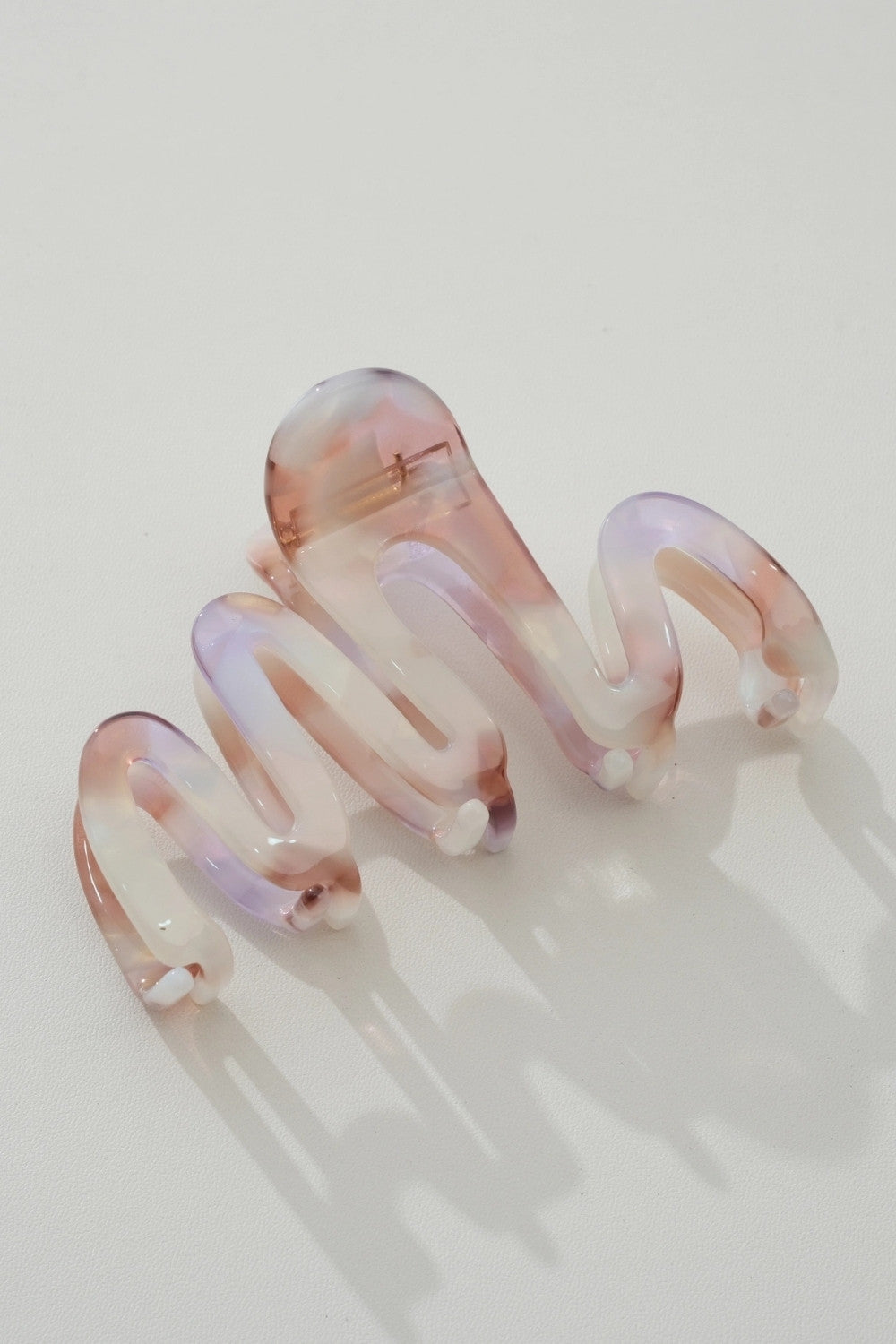 A translucent spiral hair claw clip in a swirled pink, white, and lavender acetate, displayed on a clean off-white background with soft shadows.